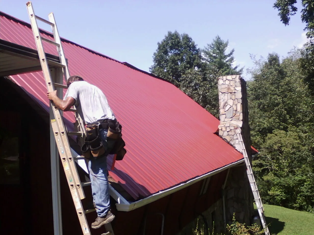Roofer on a ladder beside a red metal roof and stone chimney — Soffit & Fascia Repair by Roofing Brothers in Queens Village